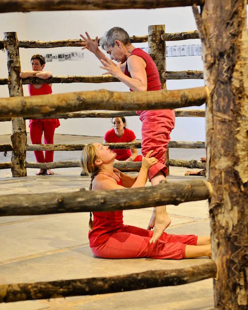 Mendl Shaw with dancers dressed in red dance in a gallery space that is enclosed by a wooden fence